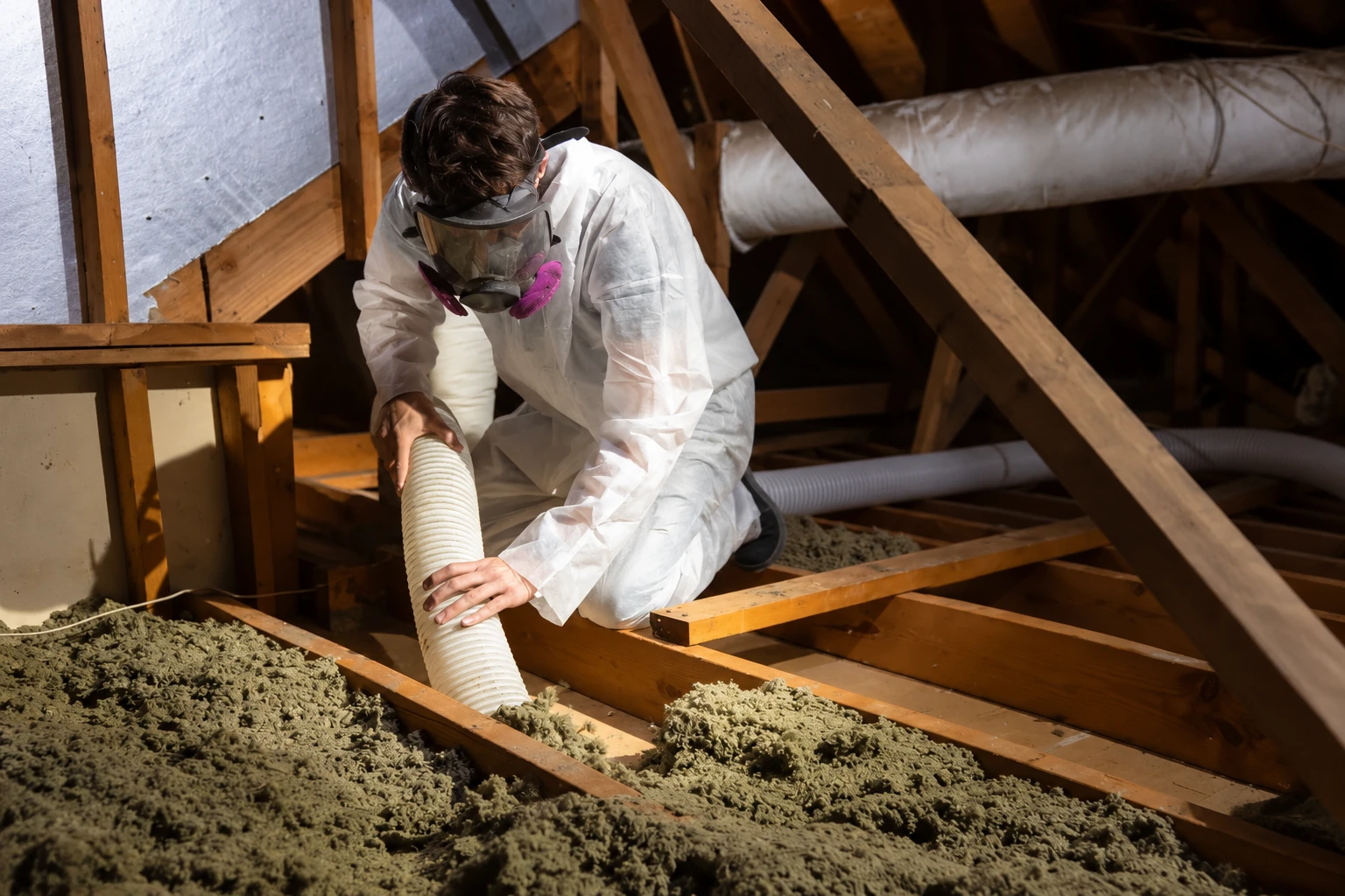A worker in protective coveralls and a respirator guides a vacuum hose over loose attic insulation.